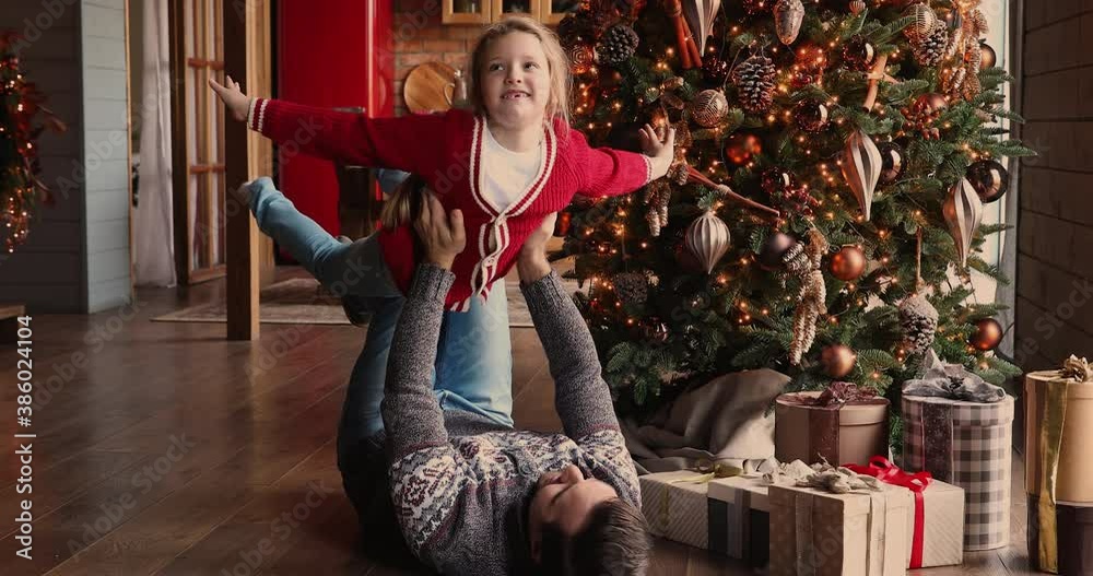 Strong dad lying on wooden floor at decorated house hall on Christmas Eve play with small daughter lifts up child stretched arms imitates plane, imagines flying in air. Family travel, xmas fun concept