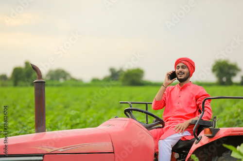 young indian farmer sitting on tractor and talking on smartphone at field