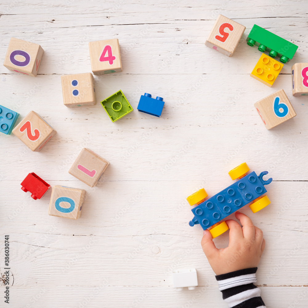 Obraz premium Development of multi-colored cubes and bricks on a white wooden table. Wooden cubes with different numbers. Top view of the kid's hands with a blue toy car and other educational toys.
