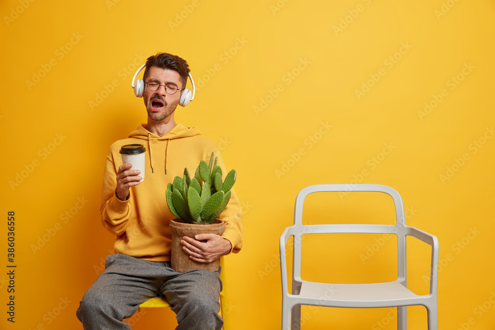 Φωτογραφία Sleepy bored young hipster man spends weekend at home drinks refreshing coffee and poses on chair with cactus listens favorite music in headphones isolated over yellow background