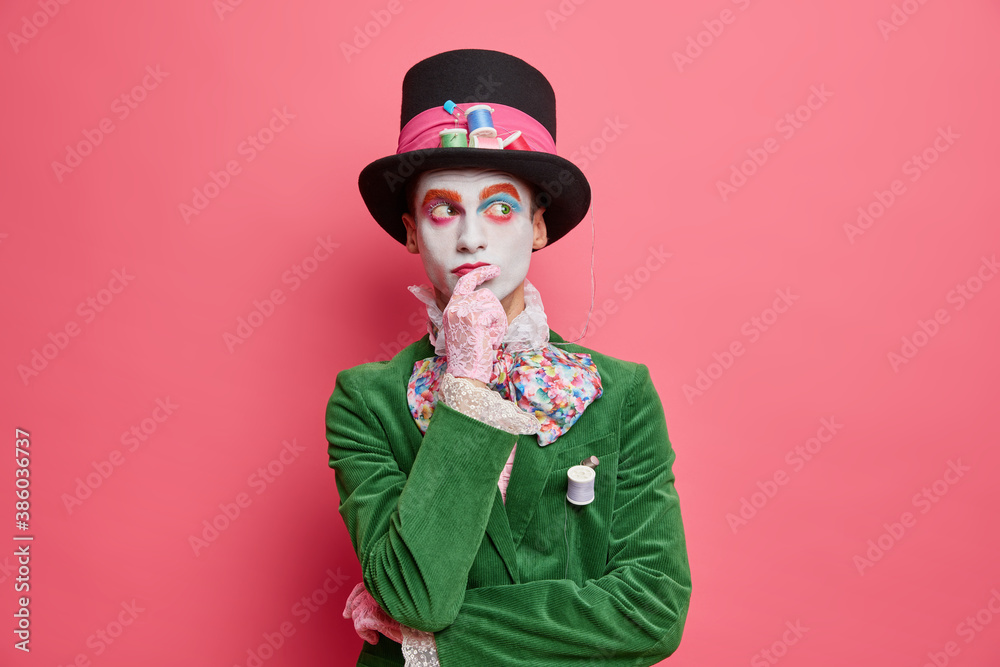 Φωτογραφία Thoughtful holiday performer thinks how to entertain people on party dressed in hatter costume wears colorful makeup poses pensive against rosy background