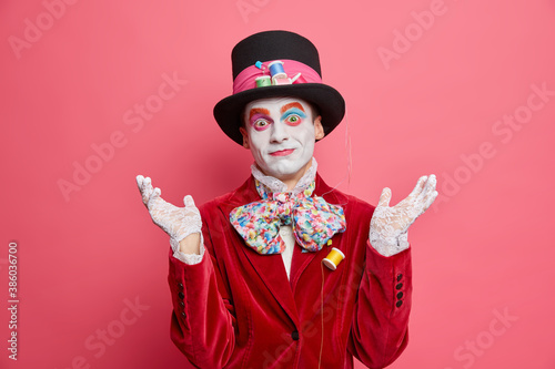 Puzzled man hesitates whether to have mad tea party raises hands in clueless gesture comes on festival wears costume and hat poses against pink background. Street performer dresses for halloween