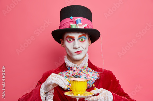 Horizontal shot of serious male hatter poses with cup of tea wears hat has manners of aristocratic gentleman dresses for masquerade carnival poses indoor. Halloween and entertainment concept photo