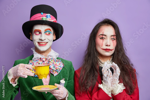 Shot of friendly positive hatter with colorful makeup enjoys tea party serious spooky woman has bloody scars keeps hands with intention to do something. Scary couple on halloween. Secret of magic