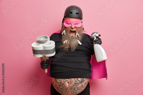 Super cleaning concept. Funny bearded janitor ready to clean up room holds plunger and detergent wears costume exclaims loudly poses against pink background. Housekeeping cleanliness hygiene concept photo