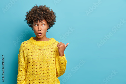 Lovely surprised Afro American woman shocked to hear unexpected relevation points thumb away and wears casual jumper impressed by incredible thing poses against blue background. Wow looks there photo