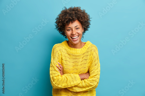 Happy joyful dark skinned woman laughs happily keeps arms folded and expresses positive emotions grins from happiness dressed in casual jumper isolated on blue background has fun or hears funny joke