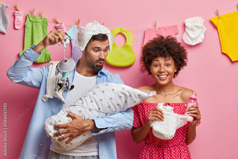 Caring affectionate father plays with newborn baby shows mobile and tries to sooth cheerful mother holds diaper and feeding bottle. Happy parents being responsible of taking care about infant child Caring affectionate father plays with newborn baby shows mobile and tries to sooth cheerful mother holds diaper and feeding bottle. Happy parents being responsible of taking care about infant child
