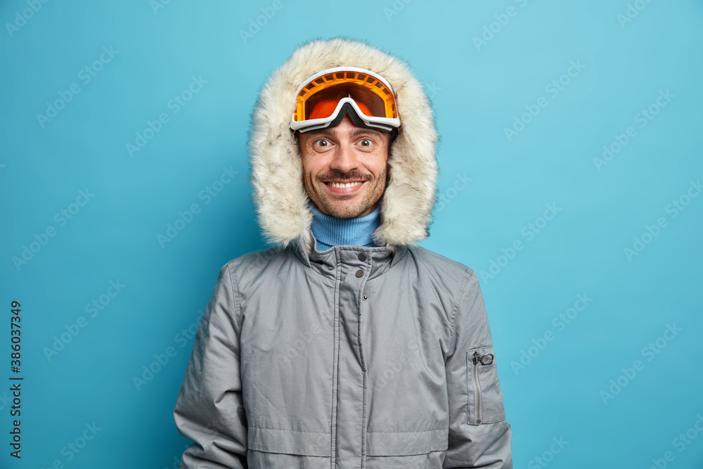 Φωτογραφία Sporty glad man enjoys winter sport recreation smiles gladfully wears ski goggles and grey jacket isolated on blue