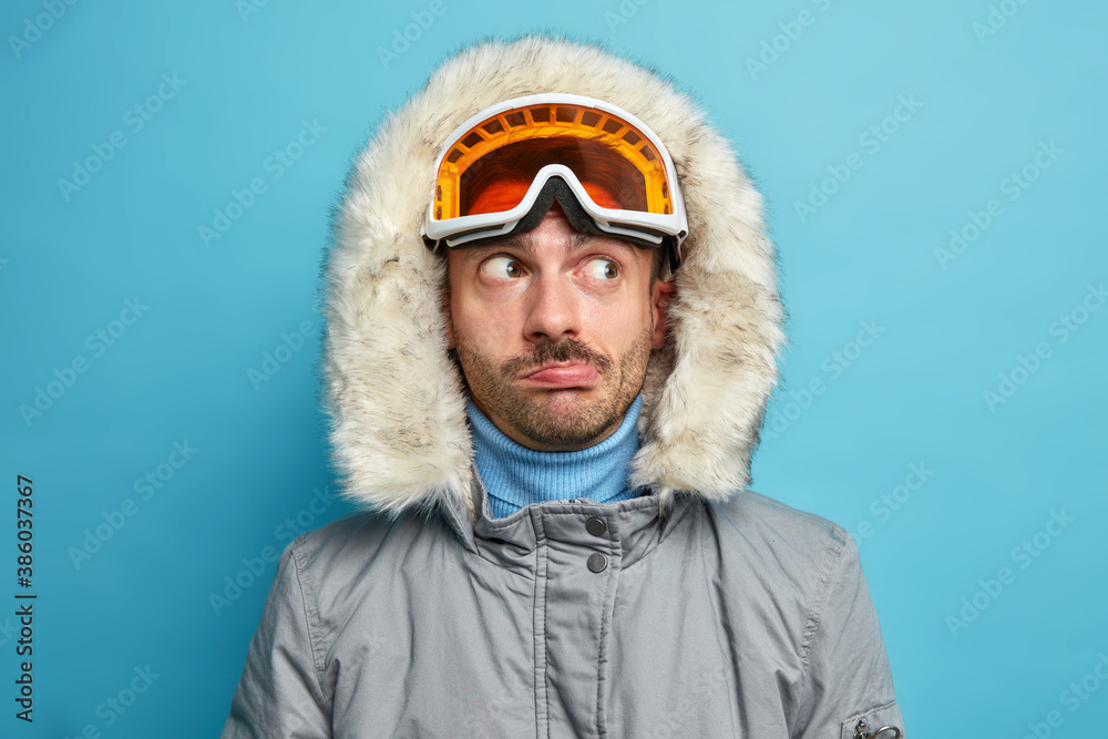 Close up shot of man hiker looks thoughtfully aside wears ski goggles and winter jacket with hood has active rest during vacation enjoys favorite sport isolated on blue background. Skier in mountains Close up shot of man hiker looks thoughtfully aside wears ski goggles and winter jacket with hood has active rest during vacation enjoys favorite sport isolated on blue background. Skier in mountains