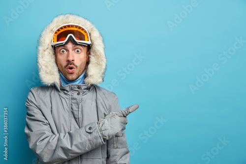 Active stupefied man trains in winter mountains goes snowboarding wears grey jacket and ski goggles indicates with surprised expression on empty space isolated over blue background. Emotional skier