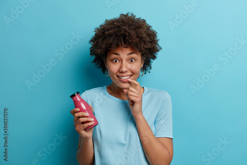 Cheerful beautiful woman with Afro hair holds fruit smoothie shake in glass bottle keeps to healthy diet dressed in casual t shirt poses against blue background. People lifestyle and nutrition concept