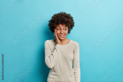 Studio shot of lovely thoughtful young African American woman bites lips and looks away with pensive expression thinks about way out wears long sleeved casual jumper isolated on blue background