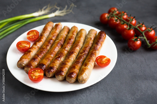 Grilled, roasted, delicious sausages on a white plate on a gray concrete background with green onion and cherry tomatos.
