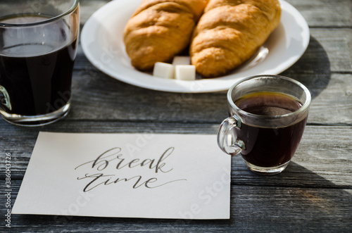 Still life with cup of coffee and croissant on the wooden background. Old retro camera and postcard are near the cup.