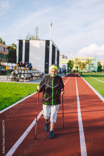 Active rest of the elderly theme. Sports and health in retirement. Caucasian very old woman with deep wrinkles doing Nordic walking exercises with sticks outside in the city stadium