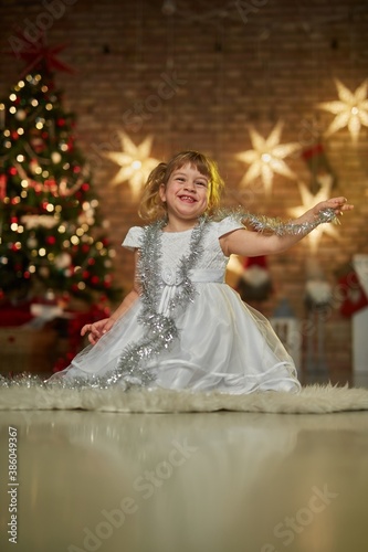 Children portrait for Christmas - little girl in white dress in front of Christmas tree at home. Christmas lights in the background. 