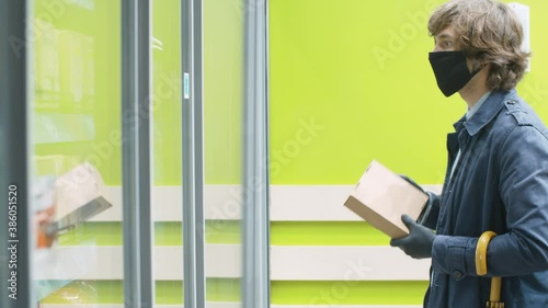 A man wearing a protective mask and gloves pulls out a cardboard box of groceries from the refrigerator in a supermarket. Protection against a viral epidemic.