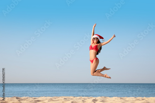 Beautiful young woman in Santa hat and bikini having fun on beach, space for text. Christmas vacation