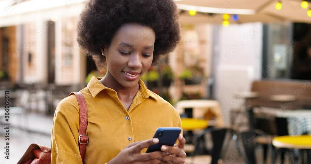 Close up of happy beautiful African American woman texting on mobile phone while standing outdoor. Joyful young female in good mood typing on smartphone on street in city. Leisure concept