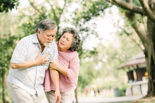 Senior Asian man holding his chest and feeling pain suffering from heart attack while his wife giving support and help outdoor at the park