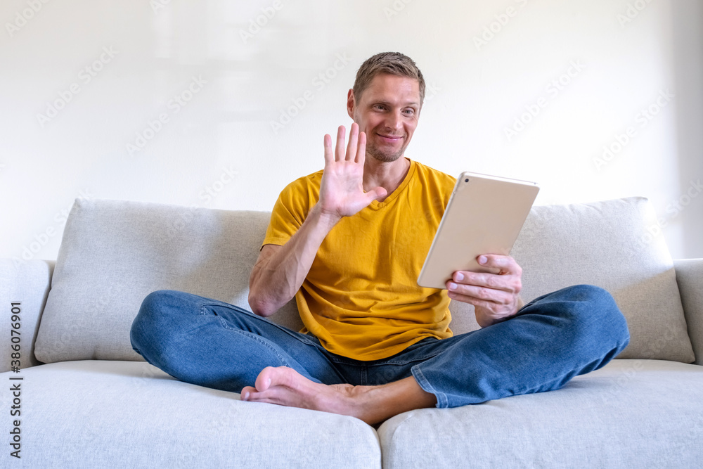 Handsome smiling man using tablet for video calling while relaxing on the couch. Guy chatting online with friends and family