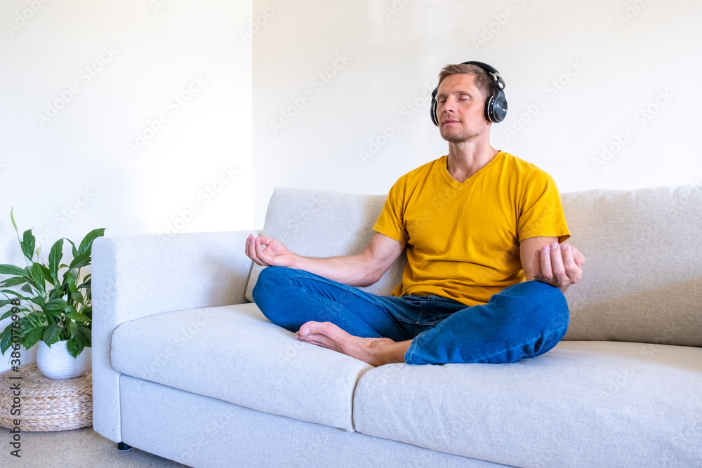 Handsome man with closed eyes sits in headphones on sofa in lotus position and meditates at home Handsome man with closed eyes sits in headphones on sofa in lotus position and meditates at home