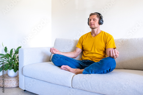Handsome man with closed eyes sits in headphones on sofa in lotus position and meditates at home