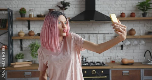 Pretty young trans woman with stylish pink wig makes selfie with contemporary smartphone posing in decorated kitchen at home photo