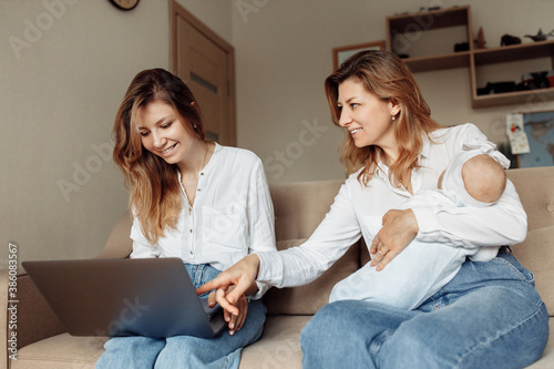 Middle aged woman with her adult daughter sit on the sofa, watching funny videos or photos, smiling, little baby girl sleep in arms of loving grandmother, three generations concept