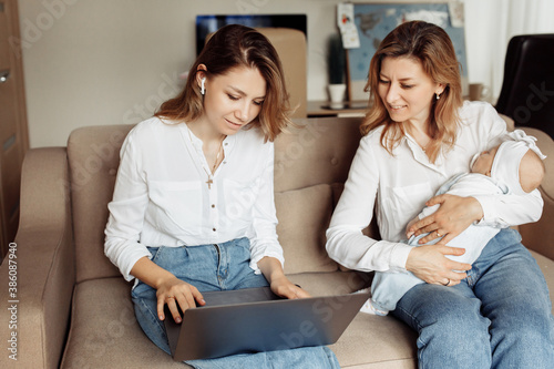 Focused woman sit at the couch, looking at the laptop, watching webinar training, young mother working at home online, while caring grandmother babysit with newborn baby girl
