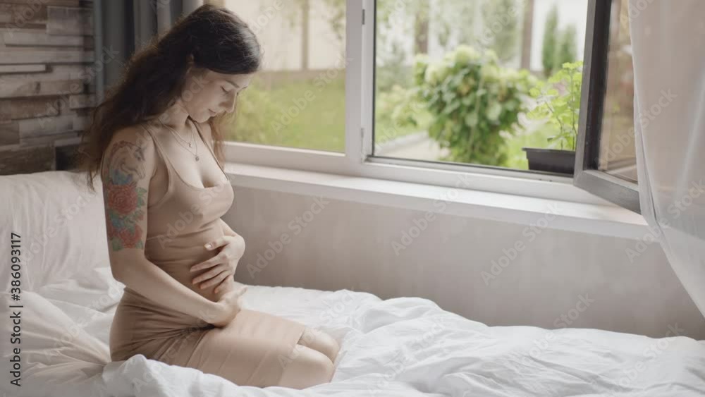 Pretty expectant mother sitting in light room. Happy pregnant woman stroking belly near window indoors. Future mom petting her stomach. Pregnant woman smiling to unborn baby Pretty expectant mother sitting in light room. Happy pregnant woman stroking belly near window indoors. Future mom petting her stomach. Pregnant woman smiling to unborn baby