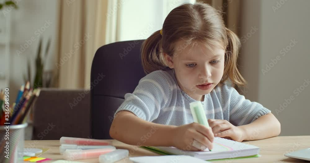 Cute little girl child 5 years old concentrating draw on paper with colored marker sitting at table at home or in kindergarten school, little daughter engaged in creativity art, talking to camera Cute little girl child 5 years old concentrating draw on paper with colored marker sitting at table at home or in kindergarten school, little daughter engaged in creativity art, talking to camera