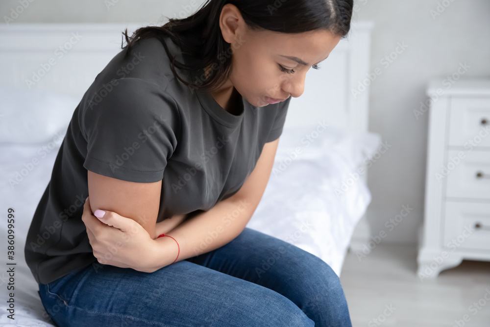 Obraz premium Sick young African American woman sit on bed at home hold belly suffer from abdominal pain or stomachache. Unhealthy biracial female struggle with painful ache, have intoxication or poisoning.