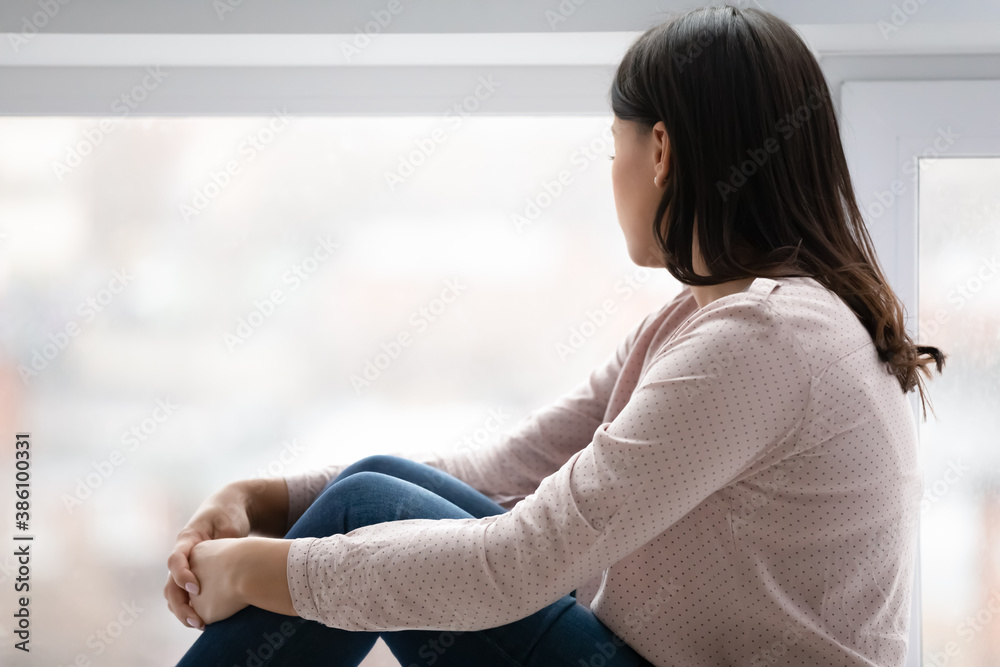 Unhappy young woman look in window distance thinking pondering, feel depressed lonely at home. Upset sad biracial female loner or outcast lack communication, suffer from depression. Solitude concept.