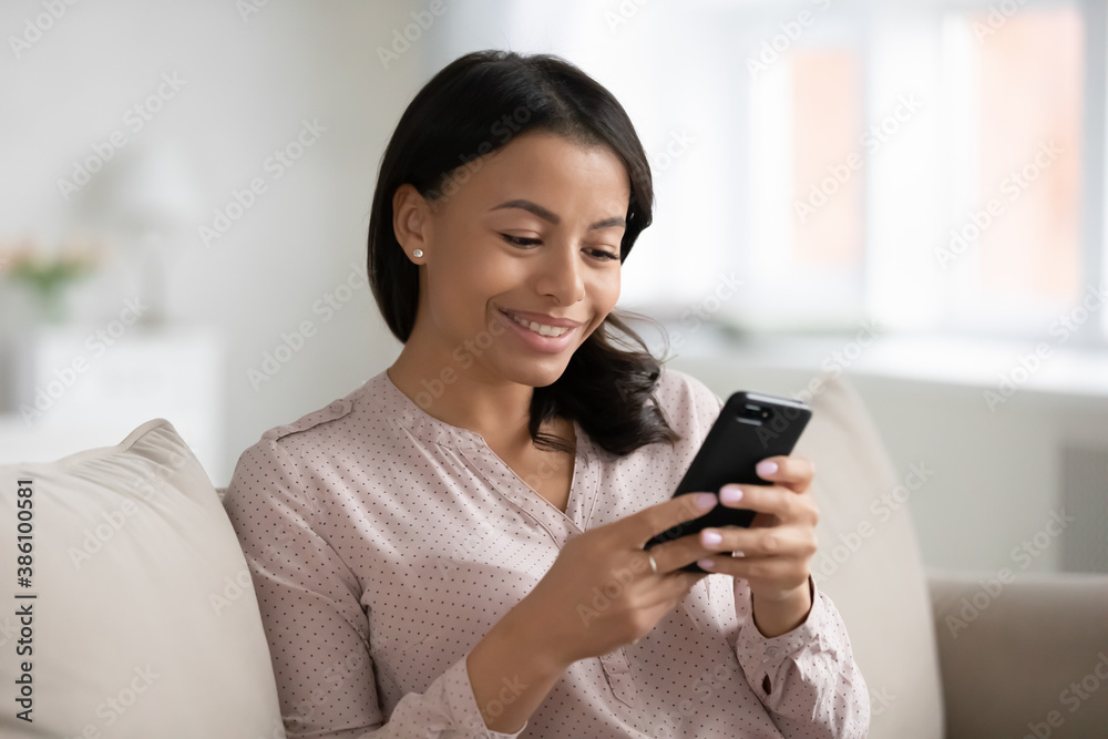 Smiling young African American woman relax on sofa in living room text message on smartphone. Happy biracial female look at cellphone screen browse surf wireless internet or shop online from home. Smiling young African American woman relax on sofa in living room text message on smartphone. Happy biracial female look at cellphone screen browse surf wireless internet or shop online from home.