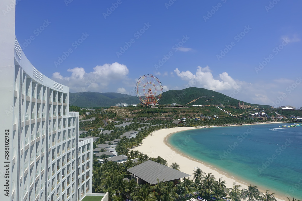 Fototapeta premium Top view of the turquoise sea, the curved line of the sandy beach, mountains. A fragment of a tall building, several villas, is visible. In the distance a colorful Ferris wheel. Vietnam. Nha Trang.