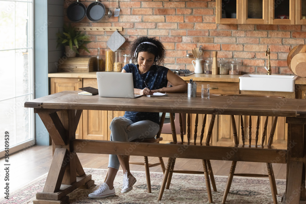 Young African American female student in headphones sit at home kitchen study online on laptop. Millennial biracial woman take distant course or training, watch webinar on computer, make notes. Young African American female student in headphones sit at home kitchen study online on laptop. Millennial biracial woman take distant course or training, watch webinar on computer, make notes.