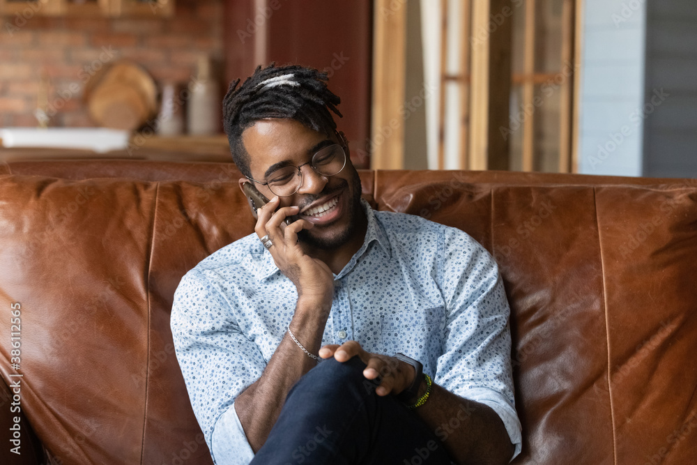 Smiling young African American man in glasses sit on couch at home have pleasant cellphone call. Happy biracial male relax on sofa in living room talk speak joke on smartphone. Communication concept. Smiling young African American man in glasses sit on couch at home have pleasant cellphone call. Happy biracial male relax on sofa in living room talk speak joke on smartphone. Communication concept.