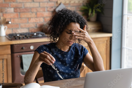 Exhausted African American young woman work on laptop at home office struggle with migraine or headache. Tired biracial female employee suffer from blurry visions or dizziness overwork on computer.