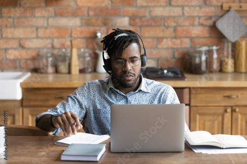 Focused biracial man in earphones sit at desk in home kitchen look at laptop screen study distant make notes. Concentrated African American male in headphones take online course training on computer.