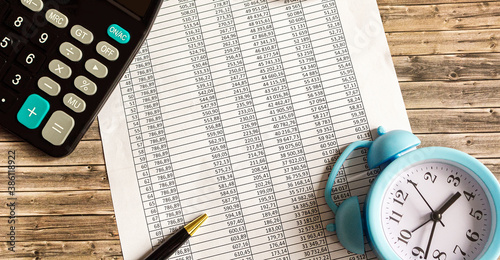 On a wooden table there is a calculator, a pen and letterheads with numbers.