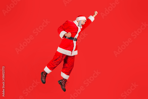 Full length cheerful positive santa claus jumping high and flying in the air like superhero, in hurry to celebrate winter holidays. Indoor studio shot isolated on red background