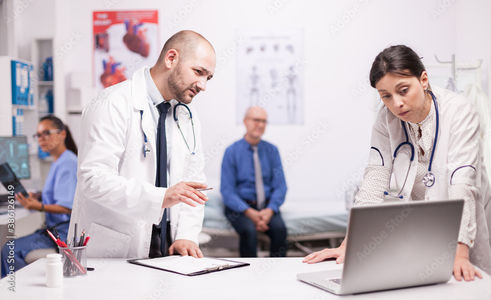 Worried doctor checking senior patient diagnosis on laptop in hospital office. Mature man sitting on bed during medical check up. Nurse holding x-ray. Worried doctor checking senior patient diagnosis on laptop in hospital office. Mature man sitting on bed during medical check up. Nurse holding x-ray.