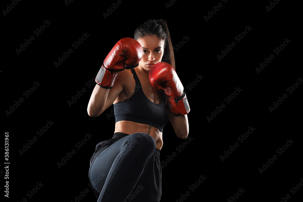 Fototapeta premium Kickboxing woman in activewear and red kickboxing gloves on black background performing a martial arts kick. Sport exercise, fitness workout.