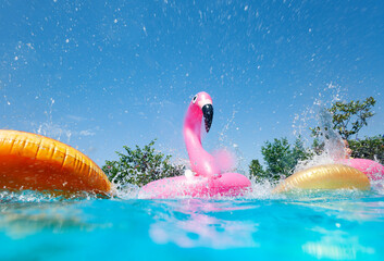 Funny action photo in the outdoor swimming pool with splashes of inflatable flamingo and doughnuts buoys rings