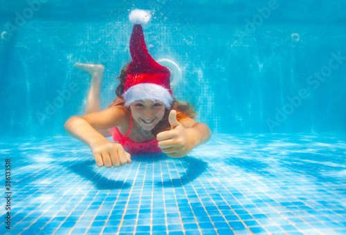 Beautiful girl with thumb up gesture dive and swim underwater wearing Santa Claus hat in the pool photo