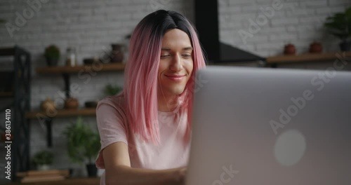 Attractive young transwoman with pink wig types message on laptop and smiles sitting at table in decorated kitchen at home photo