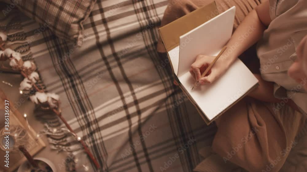 Top-view panning shot of young woman writing in diary sitting in warm and cozy room with decor lights and candles Top-view panning shot of young woman writing in diary sitting in warm and cozy room with decor lights and candles