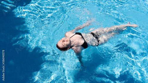 The girl swims in the pool on her back. Blue water around a floating and smiling girl in a black swimsuit.